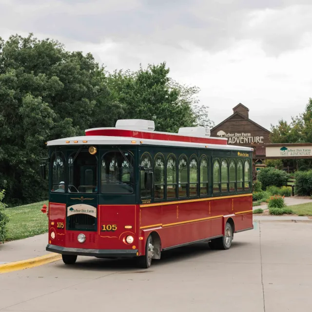 A red and green trolley bus drives past the entrance sign of Arbor Day Farm Adventure amid lush greenery and cloudy skies.