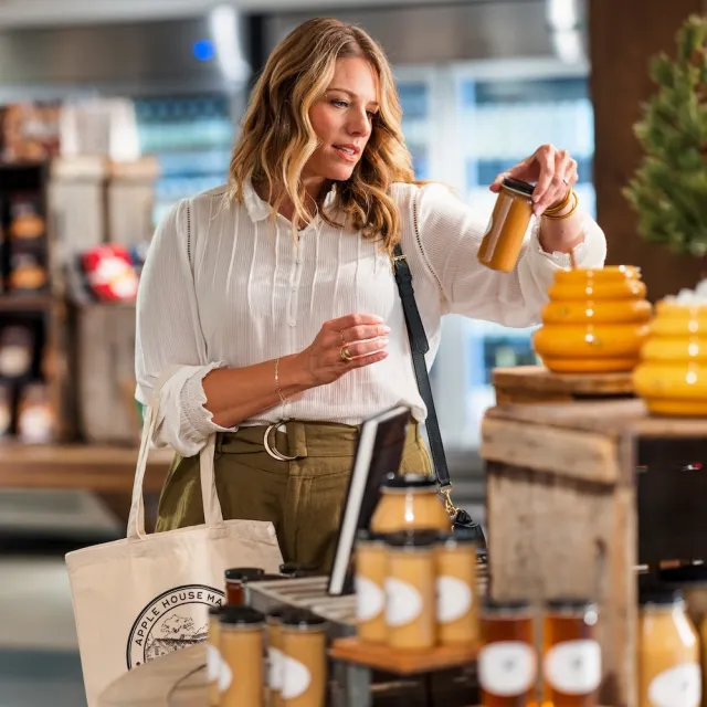 A woman inspects a jar of honey at a market stall filled with various jars and bottles of preserves and honey products.