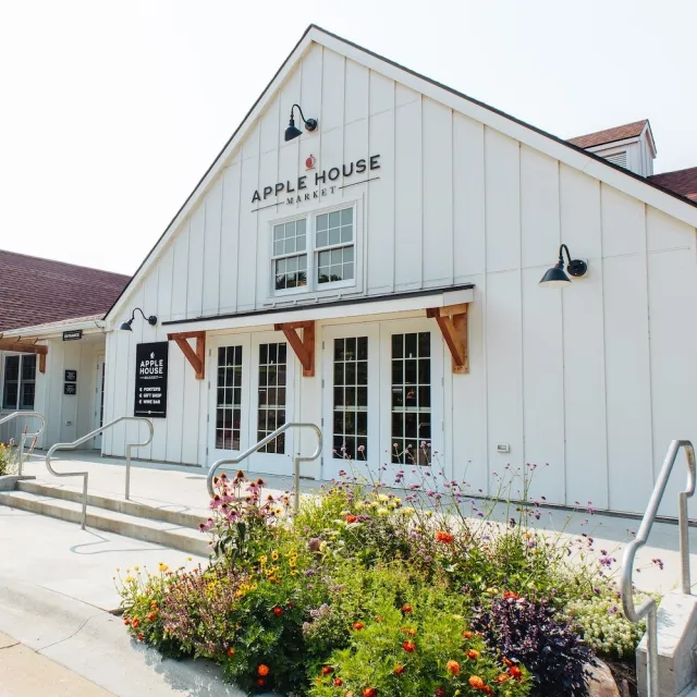 Exterior of Apple House Market featuring a large white building, colorful flower beds, and glass double doors.
