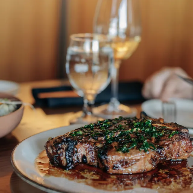 A juicy steak garnished with fresh herbs, served on a plate with wine and water glasses in a cozy dining setting.