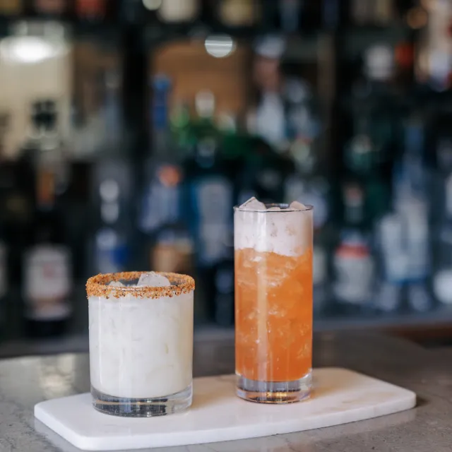 Two refreshing cocktails on a marble tray: a creamy white drink with a rim of spices and a vibrant orange drink with ice.