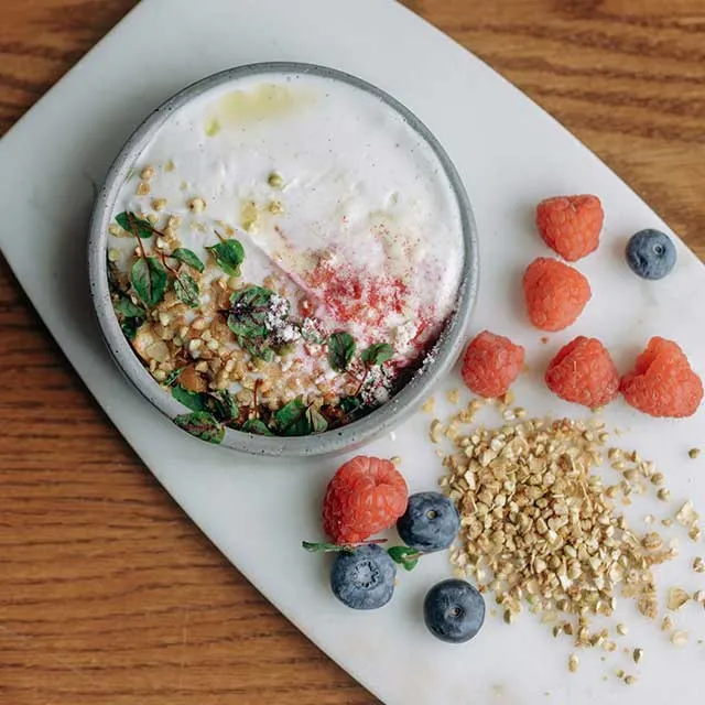 A bowl of creamy yogurt topped with granola, fresh raspberries, blueberries, and mint, on a wooden surface.