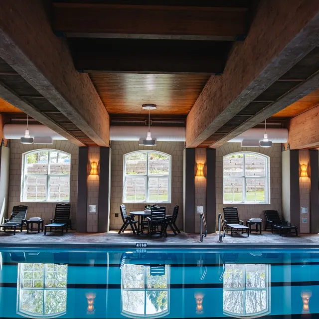Indoor swimming pool with wooden beams, lounge chairs, and large windows reflecting bright light and greenery.