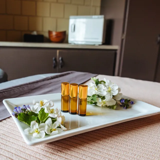 A white tray holds three amber essential oil bottles surrounded by fresh flowers and herbs, set in a tranquil spa-like room.