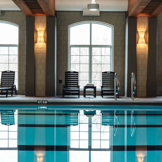 Indoor swimming pool with reflections, featuring lounge chairs, large windows, and warm lighting.
