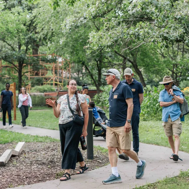 A group of people walks together on a path through a green park, surrounded by trees and plants.
