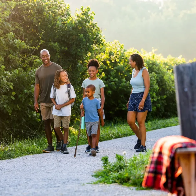 A family walks along a gravel path, surrounded by green foliage, enjoying a sunny day outdoors. A child holds a walking stick.