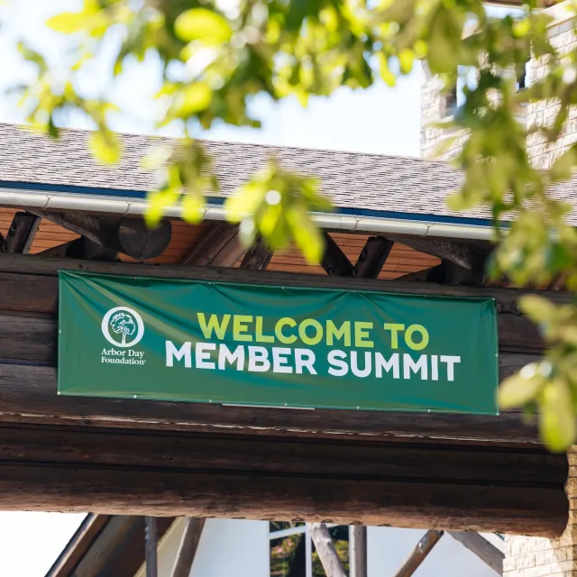 A green banner reading "Welcome to Member Summit" hangs above a rustic wooden entrance, framed by leafy branches.
