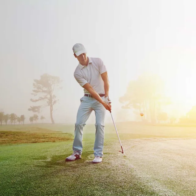 A golfer in a white polo swings his club on a misty morning, with a soft sunlight illuminating the peaceful golf course.
