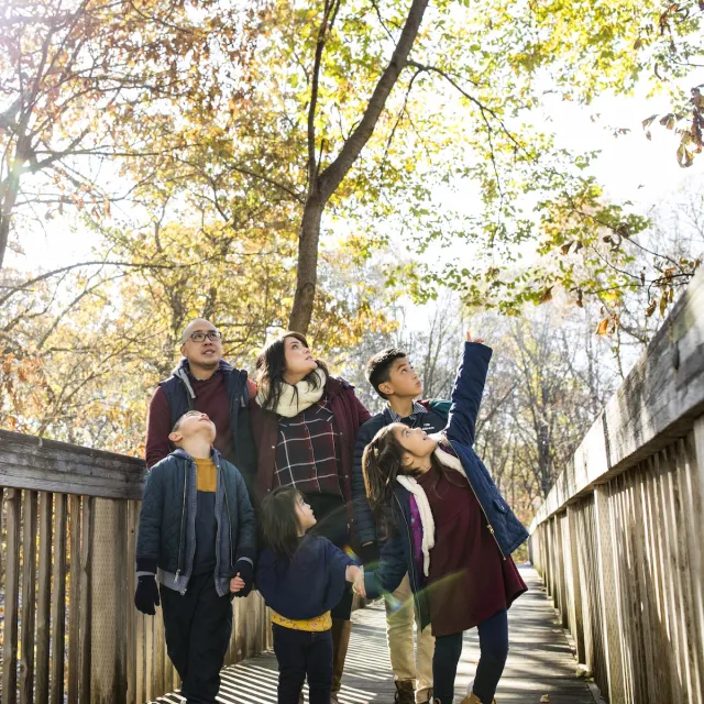A family of six, dressed casually, joyfully poses on a wooden path surrounded by autumn foliage.