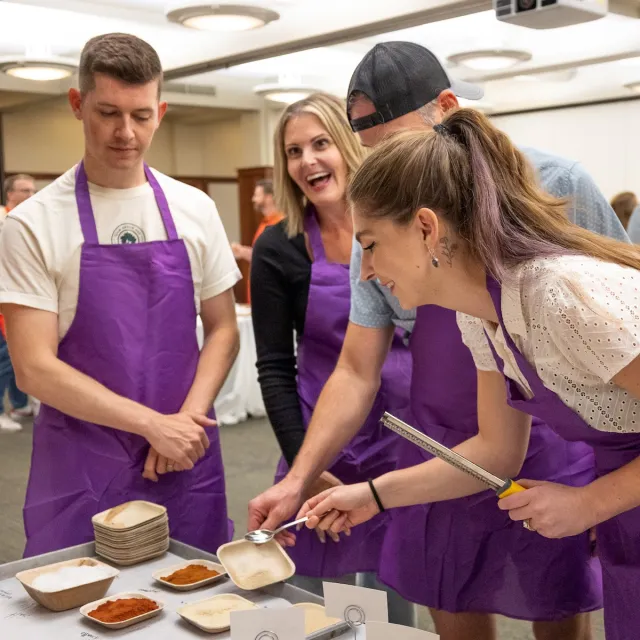 A group of people in purple aprons gather around a table filled with spice bowls, measuring ingredients for a cooking activity.