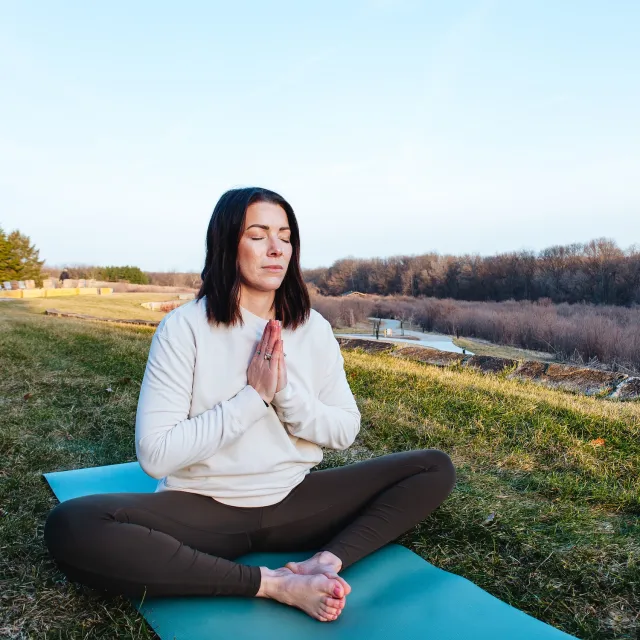 A person sits cross-legged on a yoga mat outdoors, hands in a prayer position, surrounded by grass and a scenic landscape.