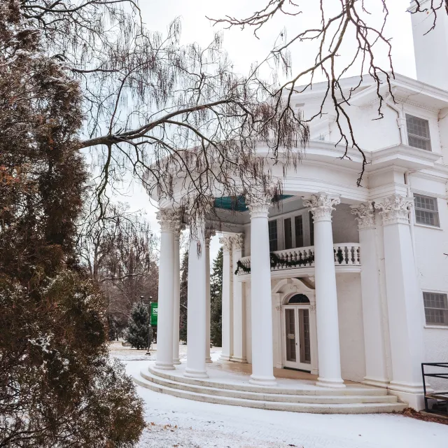 Elegant white building with tall columns and intricate details, surrounded by snow-dusted trees and branches, creating a serene winter scene.