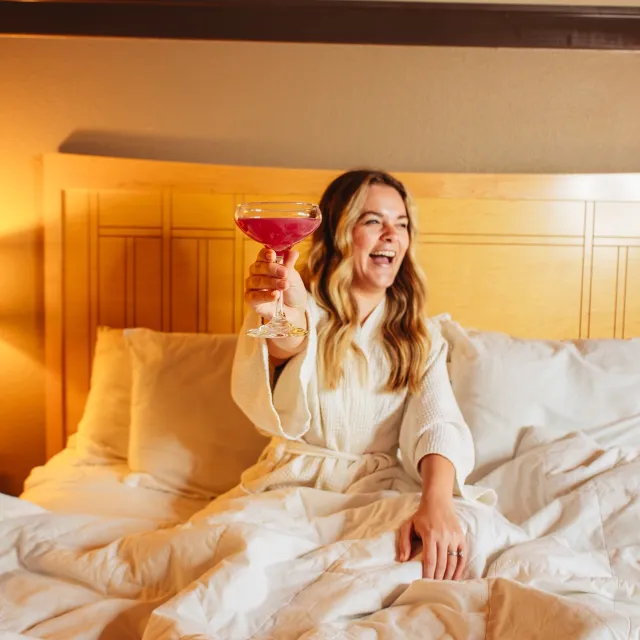 A woman in a white robe sits on a bed with white linens, joyfully raising a glass of red cocktail.