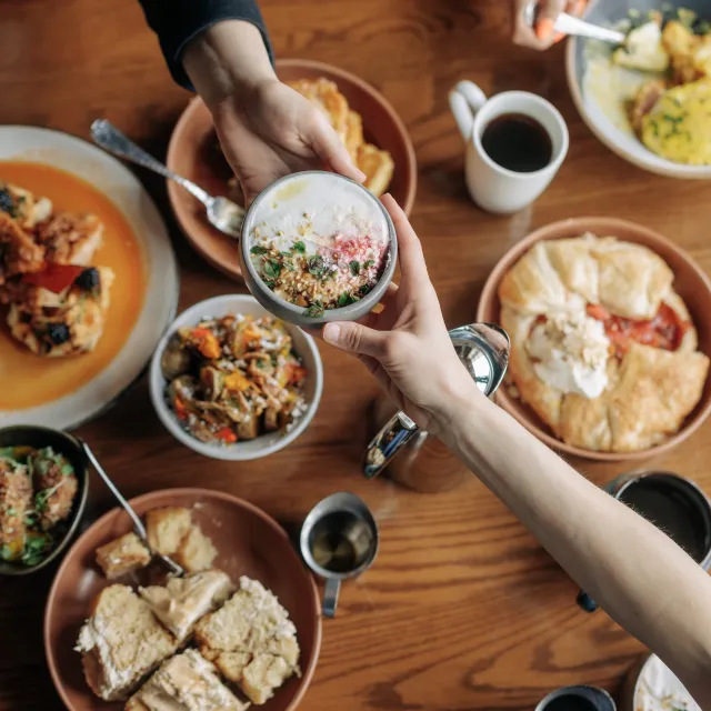 A hand holds a small bowl of colorful yogurt and toppings over a table filled with various dishes and coffee mugs.