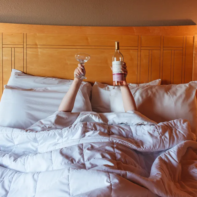A cozy bed with white sheets; two hands emerge from the covers, one holding a champagne glass, the other a bottle of rosé wine.