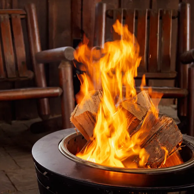 Cozy cabin scene with a vibrant fire in a metal pit, surrounded by wooden chairs.