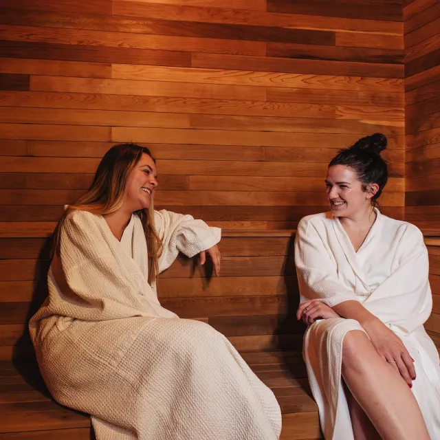 Two women in white robes relax and chat in a wooden sauna, surrounded by warm tones and a serene atmosphere.