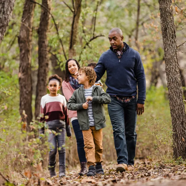 A family walks together on a forest trail, surrounded by trees and autumn foliage, enjoying a day outdoors.