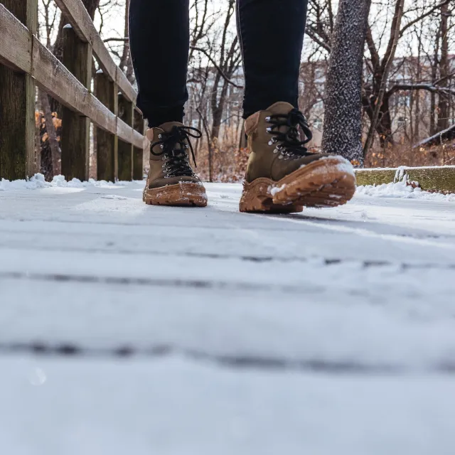 A person wearing brown boots walks on a snow-dusted wooden path in a winter forest.
