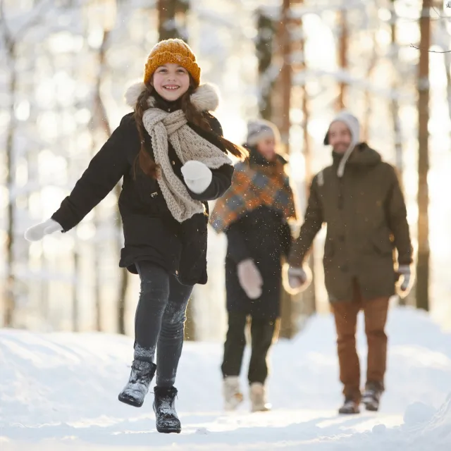 Young girl in winter clothes joyfully running on a snowy forest path, followed by two adults.