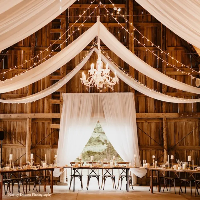 Rustic wooden barn wedding venue with dining areas with chandeliers and drapes hanging from the ceiling.
