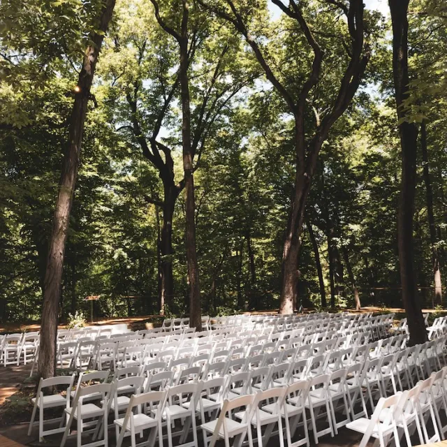 White chairs sitting in an outdoor venue location surrounded by tall green trees.
