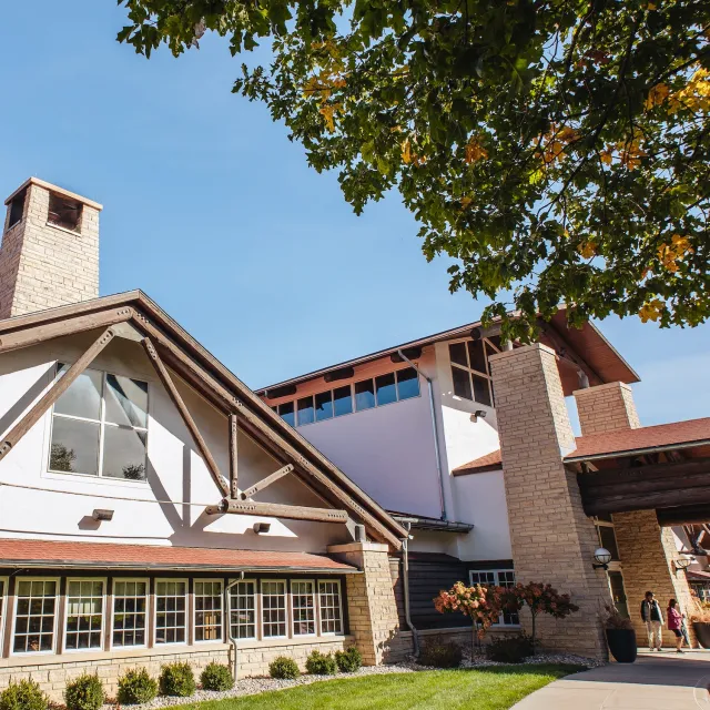 A modern building with a pitched roof, stone accents, and large windows, surrounded by greenery and a blue sky.