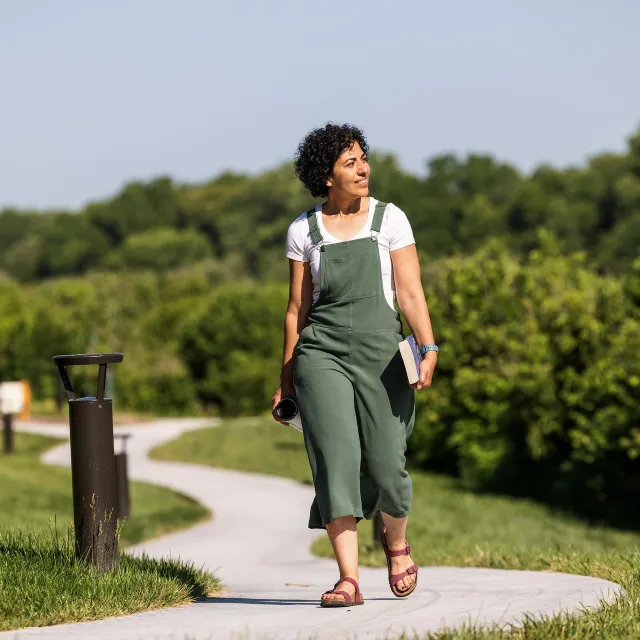 A person in a green jumpsuit walks along a curved pathway in a sunny park, holding a book and a camera. Lush greenery surrounds the path.