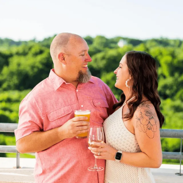 Male and female standing outside in a dress and short sleeves with drinks in their hands laughing at each other.