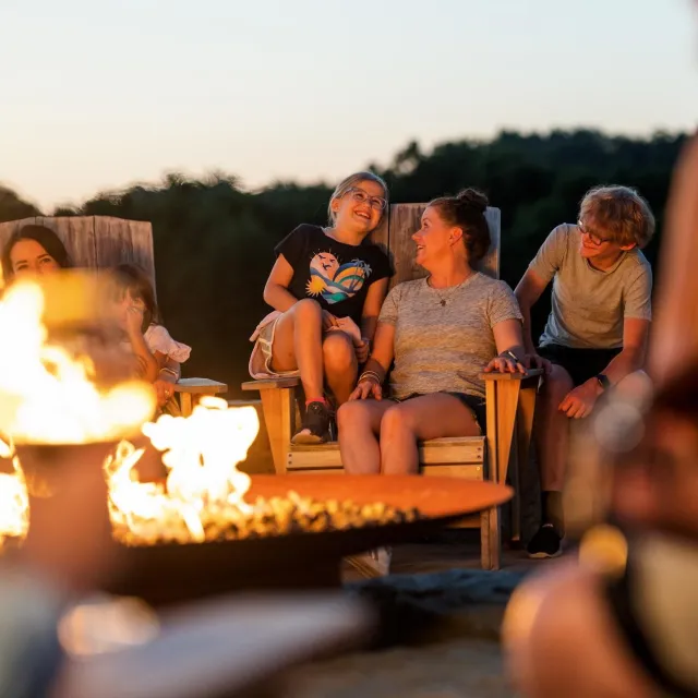 Group of people sitting around a fire pit outdoors at dusk, enjoying a relaxed evening together.