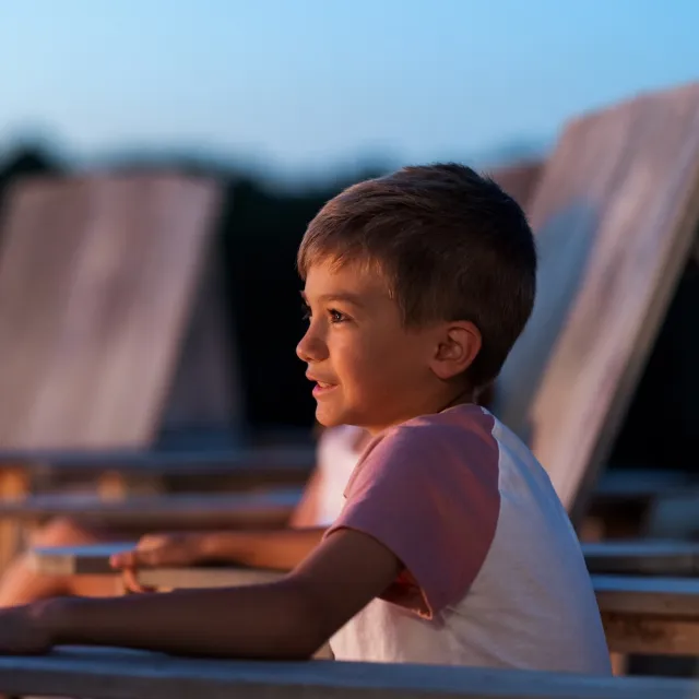 A young boy, smiling, sits in a wooden chair at sunset.