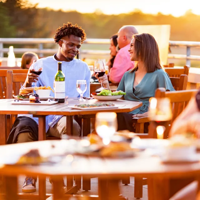 Couple enjoying outdoor dinner at sunset, raising wine glasses, with plates of food and a wine bottle on the wooden table.