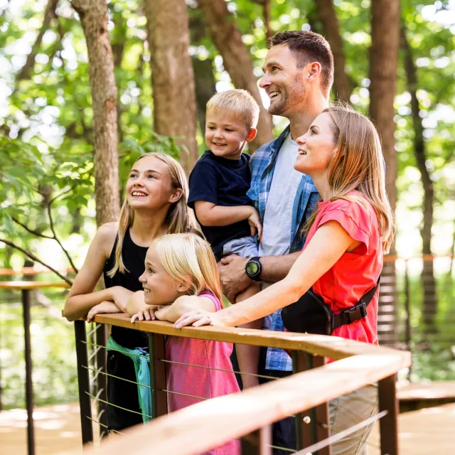 Family of five with two adults and three children standing on a wooden platform surrounded by green trees in a park.