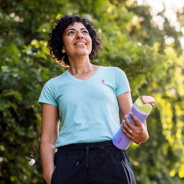 Person in a light blue shirt and black pants holding a colorful water bottle outdoors with green foliage in the background.