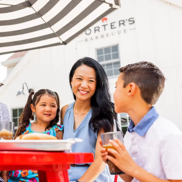 Smiling mother and two children sitting at a red outdoor table under a striped umbrella, enjoying drinks at Porter’s Barbecue.