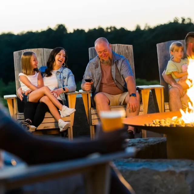 Group of people sitting on wooden chairs around a fire pit outdoors at dusk, holding drinks and enjoying a relaxed evening.