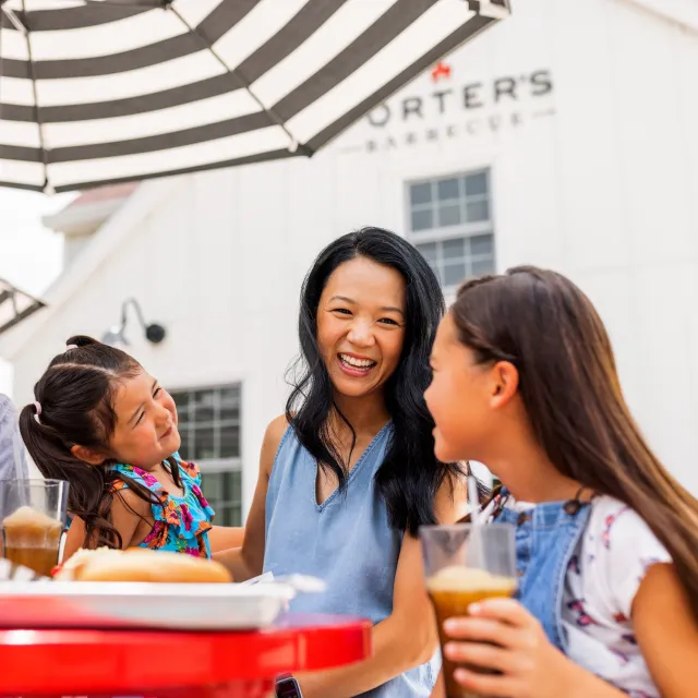 A family enjoying drinks and food outdoors at Porter's Barbecue under black and white striped umbrellas on a sunny day.