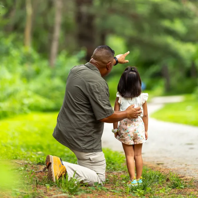 A man kneels beside a young girl on a forest path, pointing into the distance.