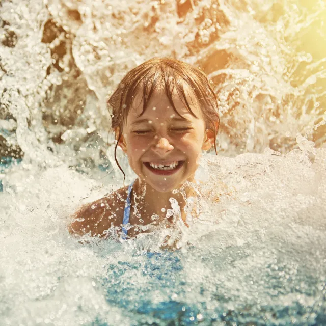 Child swimming in a pool with water splashing around, illuminated by bright sunlight.
