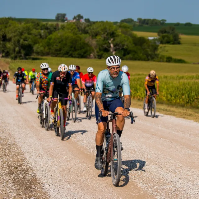 Group of cyclists riding on a gravel road through a rural landscape with green fields and trees on a sunny day.
