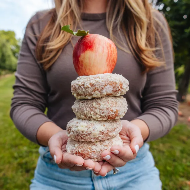 Person holding a stack of four sugar-coated donuts topped with a red apple outdoors in a green setting.