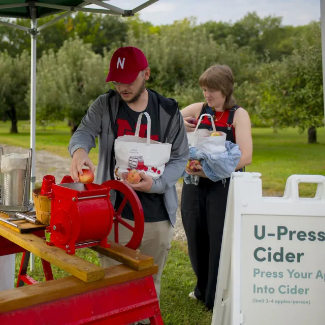Two people pressing apples into cider at an outdoor U-Press Cider station in an orchard.