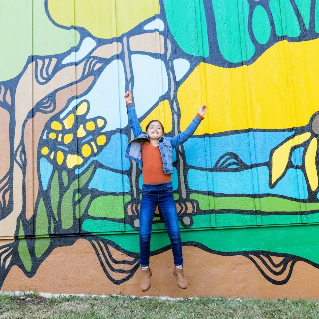 Smiling child in a denim jacket and jeans raises both arms before a bright outdoor mural of abstract plants, waves, and colorful shapes.