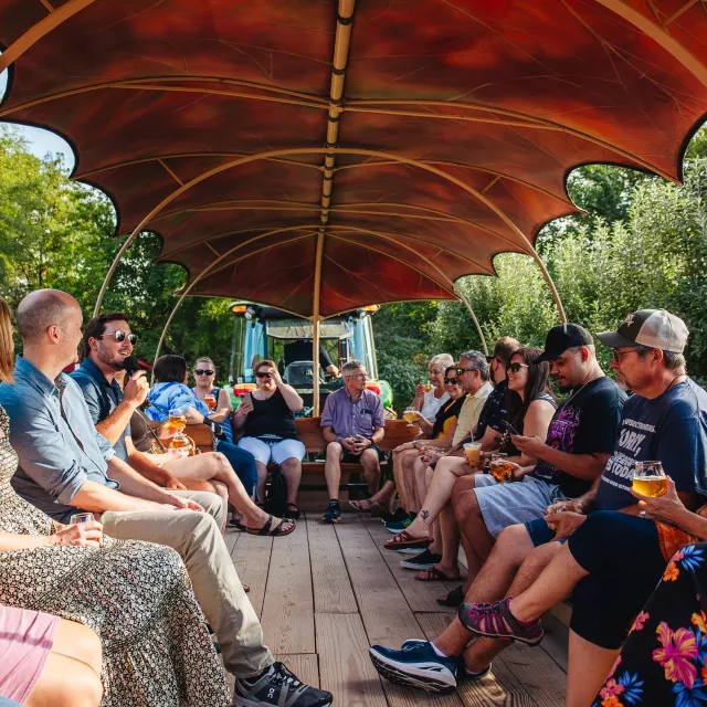 Group of people enjoying a tractor-pulled wagon ride, sitting under a canopy, drinking and socializing outdoors in a green, sunny setting.