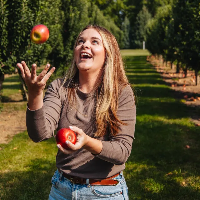 Woman with long hair juggling two red apples in a sunlit orchard between rows of apple trees on a clear day.