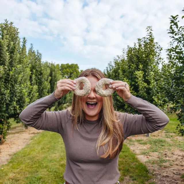 Woman holding two donuts over her eyes, smiling in an orchard with rows of green trees under a partly cloudy sky.