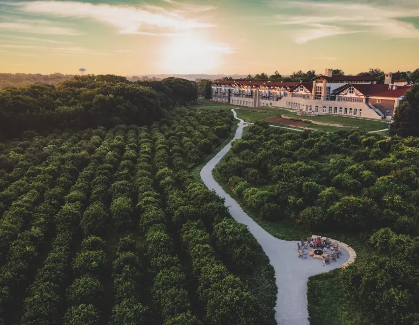 Aerial view of a serene landscape at sunset, featuring a lush green orchard, a winding path, and a large building with a red roof.