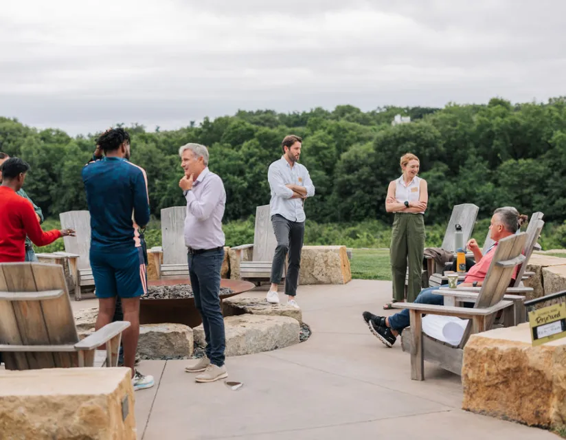 A group of people engaged in conversation around a fire pit, surrounded by nature and wooden seating in a serene outdoor setting.