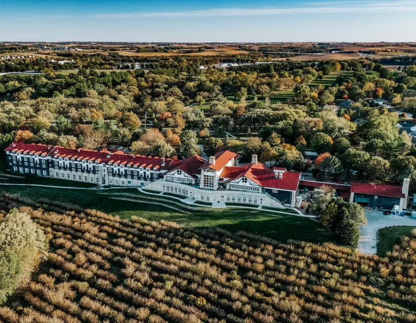 Aerial view of a historic building with red roofs surrounded by lush greenery and colorful autumn trees. Open fields in the background.